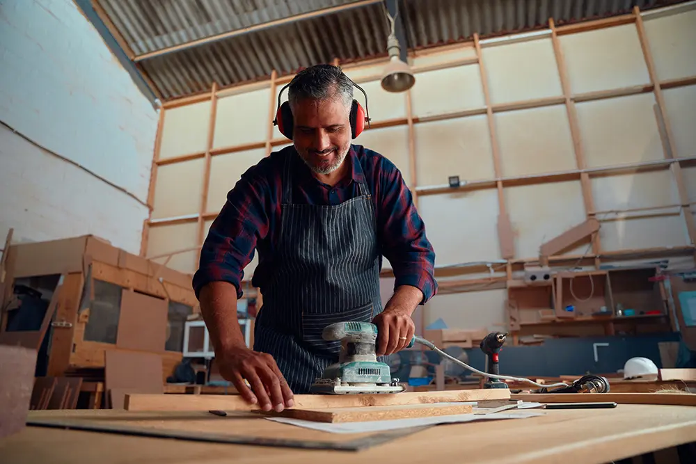 adult man smiling while wearing ear muffs and using power tool on timber in woodworking factory